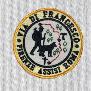 Op de afbeelding: Geborduurde ronde patch met een gouden rand. Het ontwerp toont een wandelaar met een hond, een rode lijn en de tekst "VIA DI FRANCESCO FIRENZE ASSISI ROMA". De centrale afbeelding toont een silhouet.