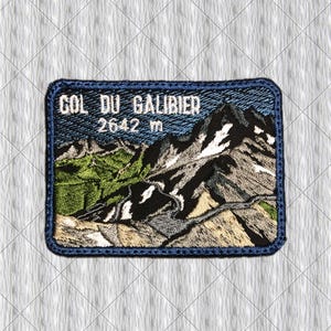 Puede incluir: Un parche bordado rectangular que muestra un paisaje de montaña con picos nevados y laderas verdes. El texto COL DU GALIBIER 2642 m está bordado en blanco en la parte superior. El parche tiene un borde azul oscuro y un fondo de tela texturizada.