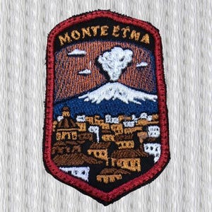 Op de afbeelding: Geborduurde patch met een landschap van de Etna, met de tekst "MONTE ETNA" bovenaan. Het ontwerp toont een uitbarstende vulkaan, een stad en een blauwe lucht. De patch heeft een rode rand.