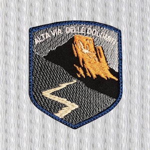 Puede incluir: Parche bordado con forma de escudo, que presenta una escena de montaña en oro y blanco sobre un fondo negro. El texto "ALTA VIA DELLE DOLOMITI" está arqueado en la parte superior, con un borde azul.