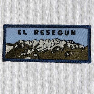 Può includere: Toppa ricamata con la scritta "EL RESEGUN" sopra una catena montuosa. La toppa ha un bordo blu, uno sfondo azzurro e la catena montuosa è raffigurata in bianco e marrone.