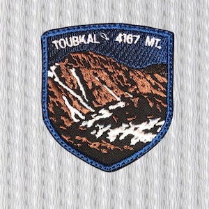 Op de afbeelding: Geborduurde patch in schildvorm met een berglandschap en de tekst "TOUBKAL 4167 MT." De patch heeft een blauwe rand. De berg is afgebeeld in bruin en wit.