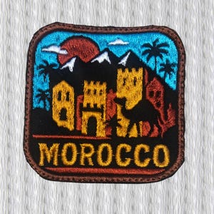 Peut inclure: Écusson brodé représentant un paysage marocain avec un coucher de soleil, des montagnes, des palmiers et des bâtiments. L'écusson a un fond noir avec le mot "MOROCCO" en lettres dorées.