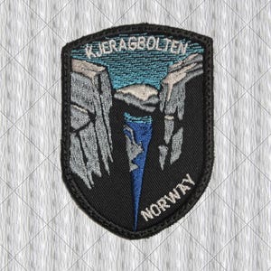 Könnte beinhalten: Gestickter Aufnäher in Schildform mit dem Text "KJERAGBOLTEN NORWAY". Das Design zeigt eine Landschaft mit grauen Klippen, blauem Himmel und einem Wasserfall. Der Aufnäher hat einen schwarzen Rand.