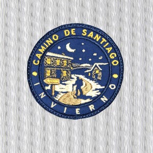 Peut inclure: Un écusson brodé bleu marine circulaire avec des lettres dorées et une scène d'hiver détaillée. Le texte indique "CAMINO DE SANTIAGO INVIERNO". L'image représente une personne marchant sur un chemin.