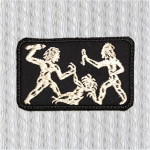 Op de afbeelding: Een rechthoekige zwarte geborduurde patch met een lichtgekleurd draadontwerp van twee staande figuren die knuppels vasthouden boven een derde figuur die op de grond tussen hen in ligt. De patch heeft een gestikte rand en een platte textuur.