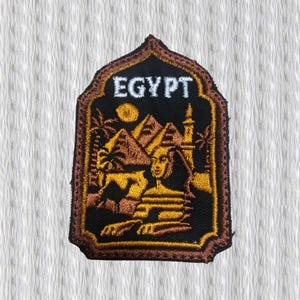 Puede incluir: Parche bordado con temática egipcia. El parche tiene un fondo negro con un borde marrón y representa pirámides, una esfinge y palmeras. La palabra "EGYPT" está bordada en blanco en la parte superior del parche.