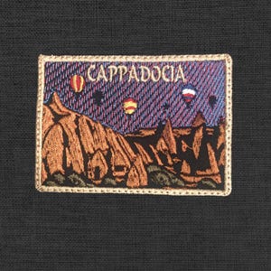 Può includere: Toppa ricamata raffigurante un paesaggio della Cappadocia con mongolfiere. La toppa rettangolare ha un bordo dorato. L'immagine mostra formazioni rocciose marroni, palloni colorati e la scritta "CAPPADOCIA" in bianco.