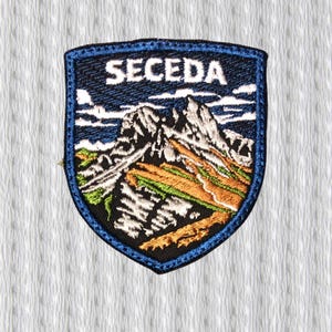May include: Embroidered patch in a shield shape, with the word "SECEDA" in white. The design depicts a mountain range with snow-covered peaks, green and brown terrain, and a blue sky. The patch is edged with a blue border.