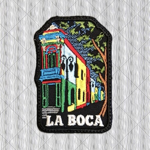 Pode incluir: Um emblema bordado retangular apresentando uma cena colorida da arquitetura de La Boca com edifícios em azul brilhante, vermelho, amarelo e verde sobre um fundo preto. Um tronco de árvore marrom está à direita e o texto LA BOCA é branco.