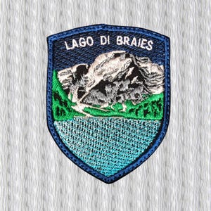 May include: A navy blue embroidered patch with the text "LAGO DI BRAIES" at the top. The patch features a mountain range, green hills, and a blue lake. The patch has a dark blue border.