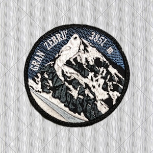 Könnte beinhalten: Ein runder gestickter Aufnäher mit einem Berggipfel, der weiße Schneeflächen und dunkelgraue Felsstrukturen zeigt. Der blaue Himmel ist mit diagonalen Linien gestickt. Der Text GRAN ZEBRU und 3851 M ist in Weiß am Rand eingestickt.