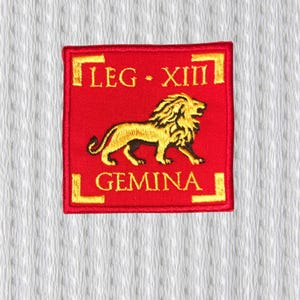 Könnte beinhalten: Ein rotes quadratisches Abzeichen mit goldfarbenen Buchstaben und einem goldenen Löwenemblem. Der Text lautet "LEG XIII GEMINA". Das Abzeichen ist mit einem goldenen Rand bestickt und vor einem weißen, strukturierten Hintergrund platziert.