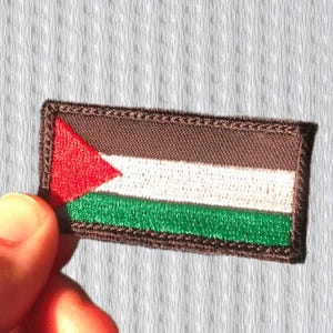 Peut inclure: Écusson brodé représentant le drapeau de la Palestine. Le patch rectangulaire a une bordure et un fond marron, avec un triangle rouge, une bande blanche et une bande verte. Le patch est tenu par les doigts d'une personne.