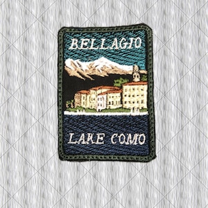Könnte beinhalten: Ein rechteckiger gestickter Aufnäher mit einer Landschaftsdarstellung von Bellagio am Comer See. Das Design zeigt weiße schneebedeckte Berge, beigefarbene Gebäude mit braunen Dächern und einen dunkelblauen See. Der Text BELLAGIO und LAKE COMO ist weiß gestickt.
