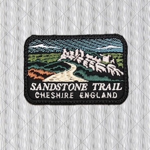 May include: A rectangular embroidered patch featuring a landscape scene with a winding path, green hills, and white rock formations under a blue sky. The black border frames the text SANDSTONE TRAIL CHESHIRE ENGLAND in white capital letters.