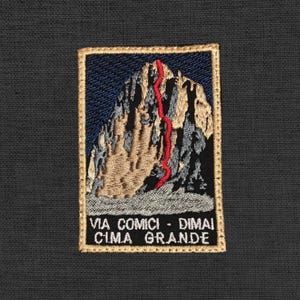 May include: Embroidered patch featuring a mountain with a red climbing route. The patch has a blue sky background and a gold border. The text "VIA COMICI - DIMAI CIMA GRANDE" is at the bottom.