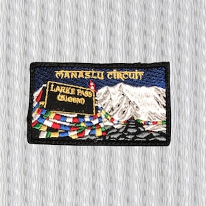 May include: Embroidered patch featuring a mountain landscape with the text "Manaslu Circuit" and "Larke Pass (5106m)". The patch has a black border, a blue sky, and colorful prayer flags.