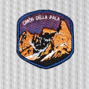 May include: Embroidered patch featuring a mountain landscape with the text "CIMON DELLA PALA" in white. The patch has a blue border and depicts orange and black mountains under a red and blue sky. The patch is on a white textured background.