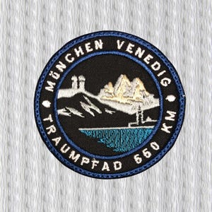 May include: Embroidered patch with a black background and a blue border. The patch features white text that reads "MÜNCHEN VENEDIG TRAUMPFAD 550 KM". The center design includes a mountain range, water, and a tower.