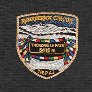 Könnte beinhalten: Gestickter Aufnäher mit dem Annapurna Circuit in Nepal. Das Design umfasst ein Gebirge, Gebetsfahnen und den Text "THORONG LA PASS 5416 m" und "NEPAL". Der Aufnäher hat einen goldenen Rand und ist vor einem dunklen Hintergrund platziert.