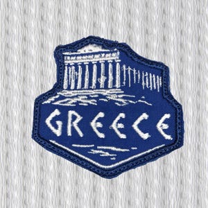 May include: A blue and white embroidered patch featuring the word "GREECE" and an illustration of a Greek temple. The patch has a shield shape with a dark blue border and is set against a white textured background.