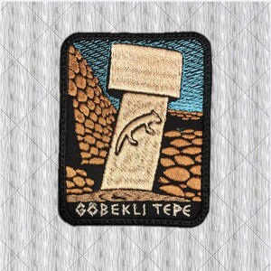 Puede incluir: Un parche bordado rectangular que muestra un pilar de piedra en forma de T de color tostado con una figura animal tallada, sobre un cielo azul y un terreno rocoso marrón. El texto GOBEKLI TEPE está bordado en blanco con borde negro.