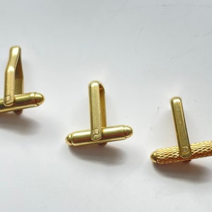 May include: Three gold cufflinks, each with a different design. The first cufflink has a simple, smooth design. The second cufflink has a slightly more intricate design with a small square in the center. The third cufflink has a textured design with a pattern of small squares.