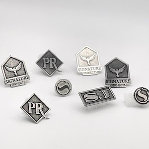 May include: A collection of eight silver metal pins with various designs. The pins feature logos, initials, and abstract shapes. The pins are all in a silver finish and have a textured surface.