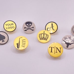 May include: Nine different round pins with various designs. The pins are yellow and gray with black designs. The designs include a skull and crossbones, a crown, a saxophone, a spade, a letter A, the number 1, a longhorn steer, and the letters TN.