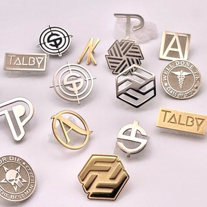 May include: A collection of various metal pins and lapel pins in silver and gold tones. Designs include letters, geometric shapes, and text such as "TALBY", "WELL DONE T.K. CLASS OF 2076", and "RIDE OR DIE THE BEST MAN".
