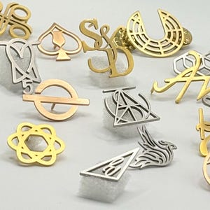 May include: A collection of various metal pins in gold and silver tones. The pins feature diverse designs, including letters, symbols, and abstract shapes. Some pins are in the shape of letters, while others are geometric or representational.