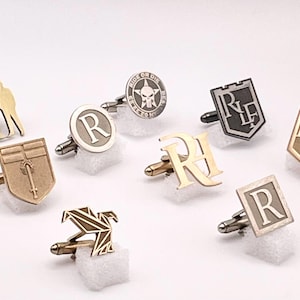 May include: A collection of nine different cufflinks featuring various designs, including a skull, a shield with a sword, a bird, and initials. The cufflinks are made of metal and have a polished finish.