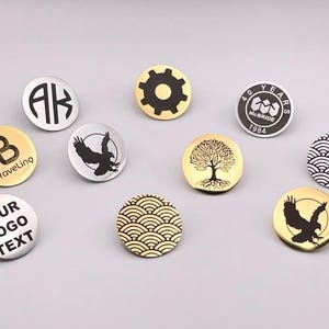 May include: Assortment of round metal pins with various designs, including initials, a gear, a tree, a bird, a wave pattern, and text. The pins are gold and silver.