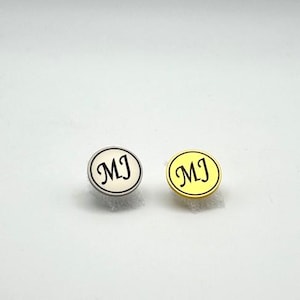 May include: Two round stud earrings. One is silver-toned with a cream-colored center and the initials "MJ" in black script. The other is gold-toned with a yellow center and the same initials.