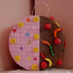 Puede incluir: Una piñata redonda, mitad rosa y mitad marrón, decorada con papel crepé. El lado rosa tiene rosetas amarillas y formas coloridas de estrellas y corazones. El lado marrón presenta bolas rojas y decoraciones coloridas en forma de gusano. Una cuerda blanca está adjunta.