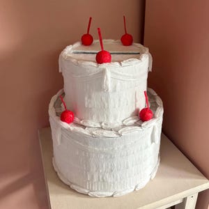 May include: A two-tiered white paper cake-shaped box with red cherry decorations. The box has a slot on top and is decorated with ruffled paper. The cake sits on a white table against a pink wall.
