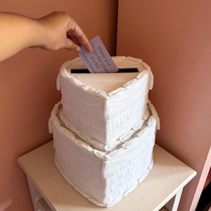 Op de afbeelding: Een witte doos in twee lagen in piñata-stijl met een gleuf aan de bovenkant. De doos is versierd met franjes. Een hand steekt een kaart in met de tekst "Thank you! Hope the piñata is a hit! - Piñata Parade."