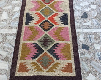 Puede incluir: Una alfombra de pasillo rectangular y colorida con un patrón geométrico. La alfombra presenta un diseño central de diamantes y zigzags en tonos de rosa, naranja, verde y marrón, enmarcado por un borde marrón oscuro y flecos.