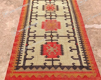 May include: Rectangular rug with a beige center and a red border. The rug features geometric patterns in black, brown, and red. The edges have a fringe detail. This rug is suitable for home decor.