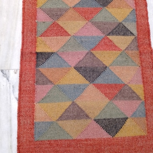 May include: A rectangular jute rug with a geometric pattern of triangles in various colours, including red, pink, yellow, and grey. The rug has a red border and fringe along the bottom edge. The rug is approximately 152 cm long.