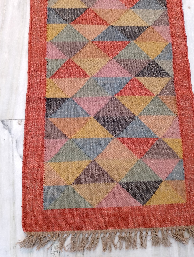 May include: A rectangular jute rug with a geometric pattern of triangles in various colours, including red, pink, yellow, and grey. The rug has a red border and fringe along the bottom edge. The rug is approximately 152 cm long.