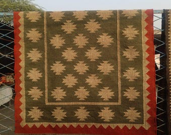May include: A rectangular rug with a geometric pattern. The rug features a dark green central field with repeating tan-coloured diamond-like shapes. A tan border frames the central field, and a red border with a zig-zag design and fringe edges the rug.