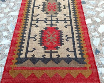 Puede incluir: Una alfombra larga y rectangular con un patrón geométrico en tonos rojos, marrones, negros y beige. La alfombra tiene flecos en el borde inferior.