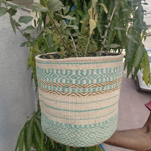 Puede incluir: Una maceta tejida con base beige y patrones geométricos turquesa y marrón. La maceta está llena de tierra y una planta verde. La maceta cuelga de una rama de árbol.