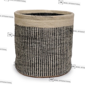 Puede incluir: Una cesta cilíndrica tejida con una parte superior beige y un patrón de rayas negras y beige. La cesta tiene una construcción robusta y una estética natural y terrosa, adecuada para almacenamiento o decoración.