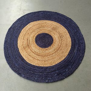 Op de afbeelding: Een ronde jute vloerkleed met een marineblauwe rand en een bruin centrum. Het vloerkleed heeft een concentrisch cirkelontwerp, met een marineblauwe ring in het bruine centrum. Gemaakt van geweven jutevezels.