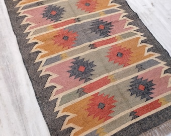 Puede incluir: Una alfombra rectangular con un patrón geométrico en tonos grises, rosa, naranja y marrón. La alfombra tiene un borde de flecos.