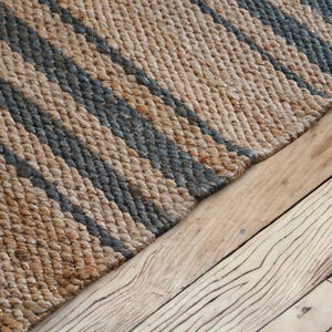 以下が含まれることがあります： 織り込まれた質感の天然繊維ラグのクローズアップ。ラグは、ベージュとグレーの色合いの斜めストライプのパターンが特徴です。ラグは、木目がはっきり見える木の床に置かれています。