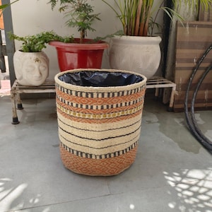 May include: Woven plant pot with a black interior lining. The pot features a geometric pattern in shades of beige, orange, and black. Several potted plants are visible in the background, including a small tree and a tall green plant.